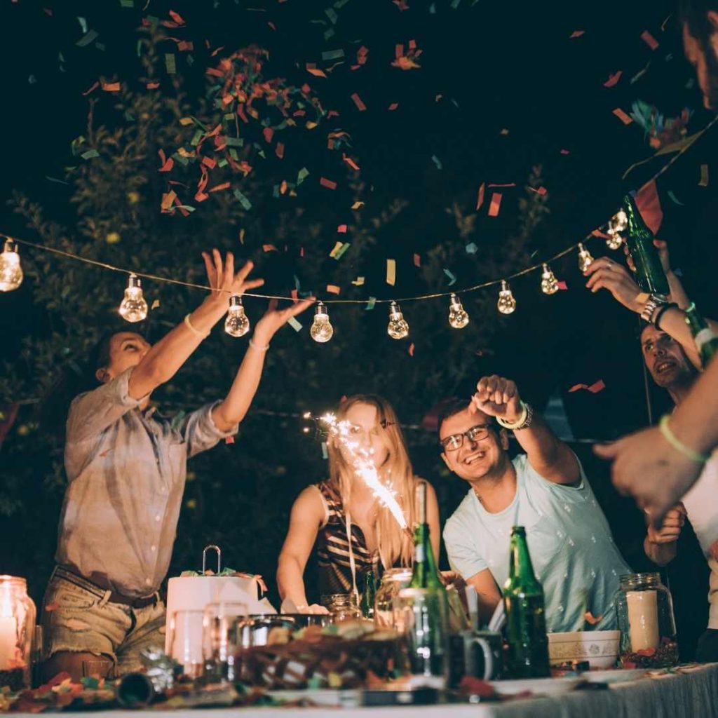 A group of people sitting at a table and having a party together with festoon lights hanging above their heads